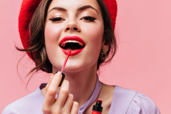 Woman in bright beret paints her lips with red lipstick. Girl in lilac blouse posing on pink background.